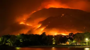 Nächtliches Waldbrand-Szenario mit flammenden Berghängen oberhalb einer dunklen Gewässer-Silhouette.