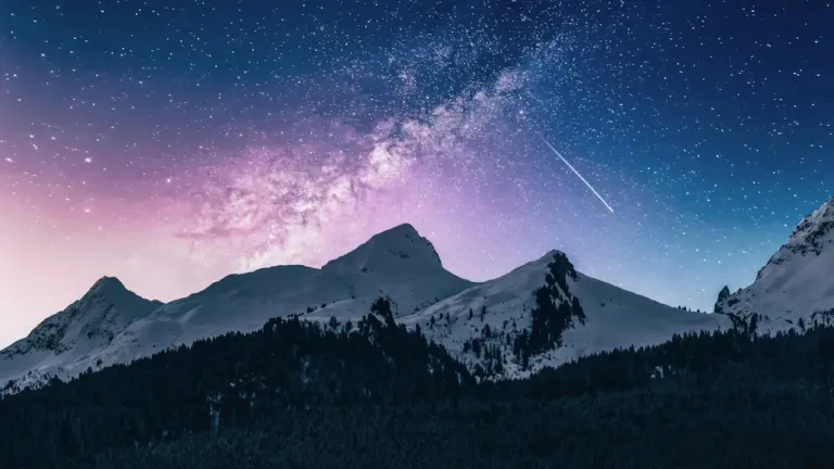 Spektakulärer Sternenhimmel mit Milchstraße und einer Sternschnuppe über verschneiten Berggipfeln in der Nacht.