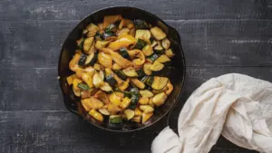Eine gusseiserne Pfanne mit gebratenen Zucchini-Stücken auf einem dunklen Holztisch.