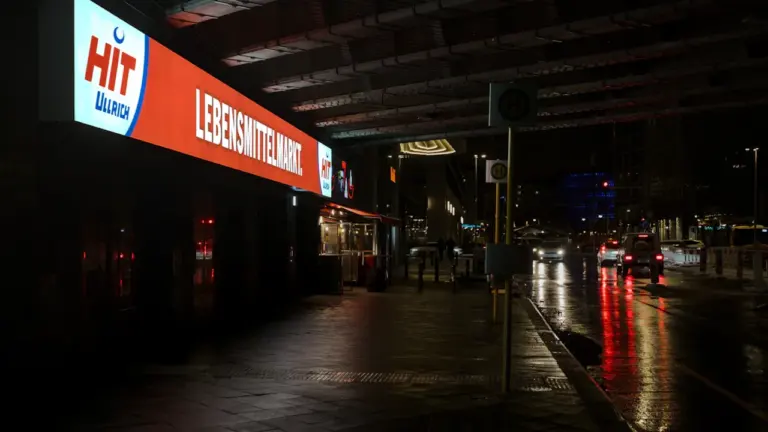 Ein beleuchtetes Schild eines deutschen Lebensmittelmarktes an einer regnerischen Straße bei Nacht.