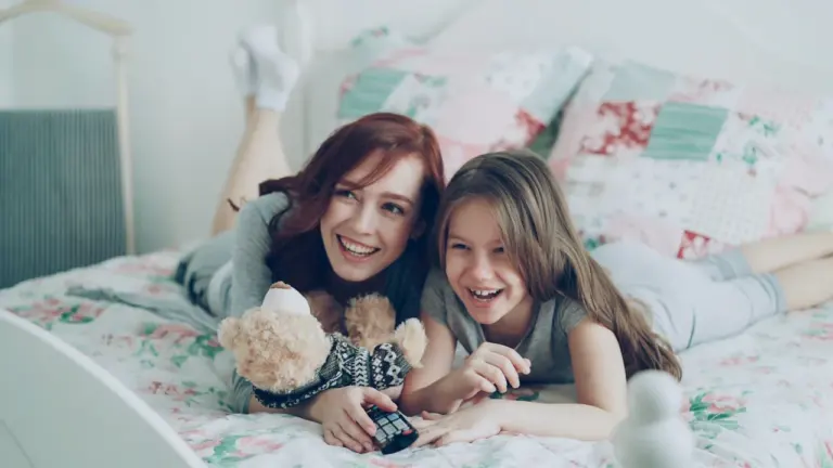 Mutter und Tochter lachen gemeinsam beim Fernsehen in einem gemütlichen Schlafzimmer am Morgen.