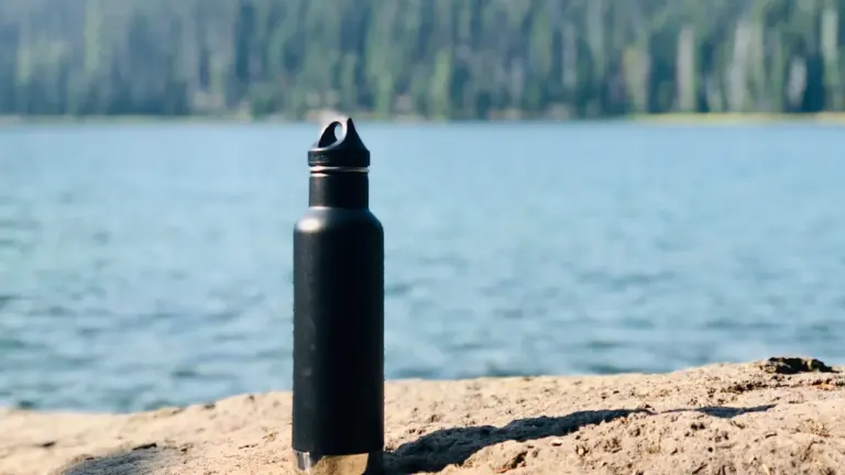 Schwarze Trinkflasche auf einem Stein vor einem See bei sonnigem Wetter.